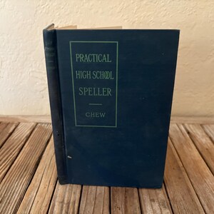 May include: A vintage navy blue book titled "Practical High School Speller" by Chew. The title is in a green rectangular box on the front cover. The book is standing upright on a wooden surface.
