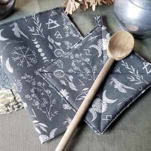 May include: Two gray fabric pot holders with white designs of moons, stars, and other symbols. A wooden spoon is lying on top of the pot holders.