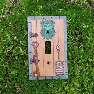 May include: A light switch cover with a green woodgrain design. The cover features a green man face, an owl, and a crescent moon. The switch is off.