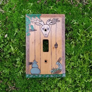 May include: A wooden light switch cover with a painted design of a deer skull, a potted plant, and a hanging lantern. The switch cover is painted in shades of brown, green, and white.
