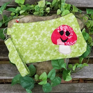 May include: A green fabric clutch with a white floral pattern and a wrist strap. The clutch features a smiling mushroom applique with a red cap and a white stem. The clutch is surrounded by green foliage.