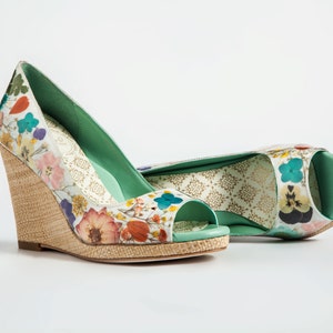 K&ouml;nnte beinhalten: Ein Paar Keilabsatzschuhe mit Blumenmuster und gr&uuml;nem Peeptoe. Die Schuhe haben einen geflochtenen Keilabsatz und einen wei&szlig;en Hintergrund mit bunten Blumen.