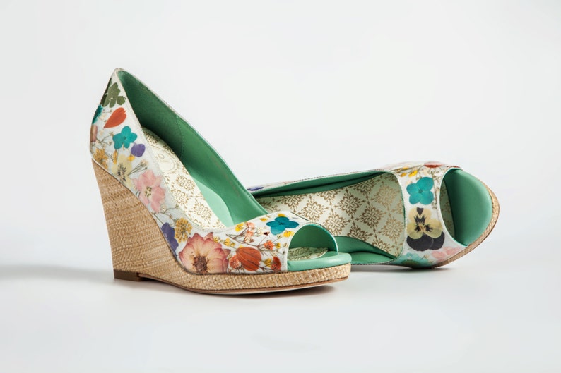 K&ouml;nnte beinhalten: Ein Paar Keilabsatzschuhe mit Blumenmuster und gr&uuml;nem Peeptoe. Die Schuhe haben einen geflochtenen Keilabsatz und einen wei&szlig;en Hintergrund mit bunten Blumen.