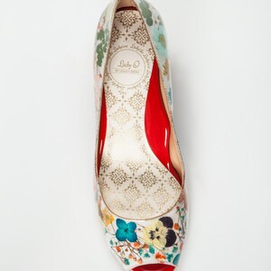 May include: A white peep-toe heeled shoe with a floral print. The shoe has a red insole and a red heel. The brand name "Lady O by Lesley Zheng" is printed on the insole.