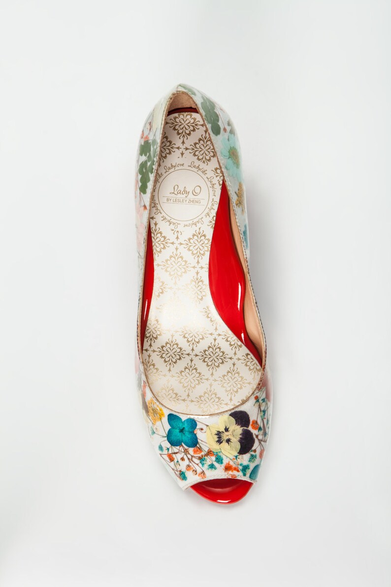 May include: A white peep-toe heeled shoe with a floral print. The shoe has a red insole and a red heel. The brand name "Lady O by Lesley Zheng" is printed on the insole.