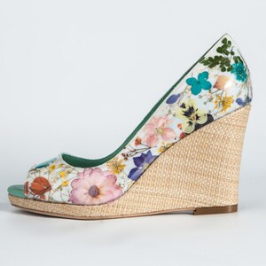 K&ouml;nnte beinhalten: Ein wei&szlig;er Keilabsatz mit Blumenmuster und einem nat&uuml;rlichen gewebten Keil. Der Schuh hat eine offene Zehenpartie und ein gr&uuml;nes Futter.