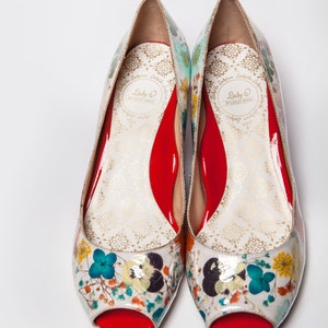 May include: A pair of white peep-toe shoes with a floral print and red soles. The shoes have a gold label that reads "Lady O by Lesley Jane."