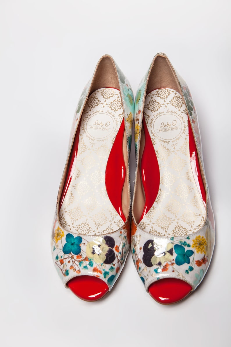 May include: A pair of white peep-toe shoes with a floral print and red soles. The shoes have a gold label that reads "Lady O by Lesley Jane."