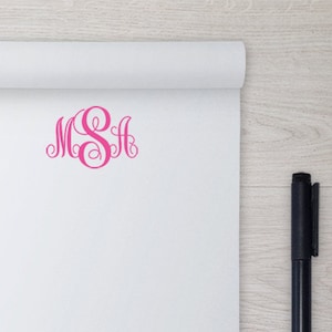 May include: A white notepad with a pink monogram "M S H" on the front. A black pen is lying on a wooden surface next to the notepad.