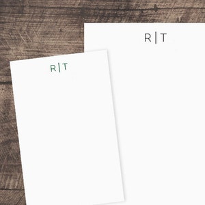 May include: Two white notepads with the letters "R|T" printed in dark green at the top of each notepad. The notepads are on a brown wooden surface.