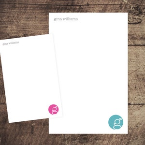 May include: Two white stationery note cards with the name "Gina Williams" printed at the top. The top card has a teal blue circle with a white letter "G" inside. The bottom card has a pink circle with a white letter "G" inside.