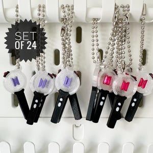 May include: A set of 24 keychains featuring a clear, round design with a logo in purple and red. Each keychain has a black handle and a silver chain. The keychains are hanging on a white pegboard.