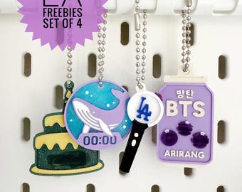 May include: A set of four hanging charms with silver ball chains. The charms include a green glittery cake, a circular purple whale design with 00:00 text, a white and blue LA light stick, and a rectangular purple BTS Arirang tag with Korean text.