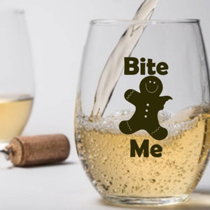Op de afbeelding: Een helder glazen wijnglas met een afbeelding van een peperkoekman en de tekst "Bite Me". Het glas is gevuld met witte wijn.