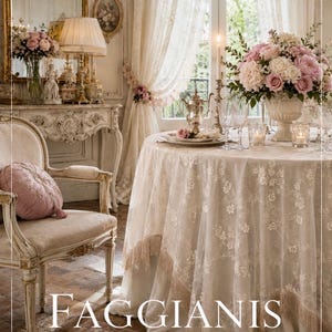 May include: A round dining table covered with a cream-colored lace tablecloth featuring floral embroidery and fringe trim. The table is set with a floral centerpiece, glassware, and a silver teapot. The text FAGGIANIS SARTORIA ITALIANA is visible.