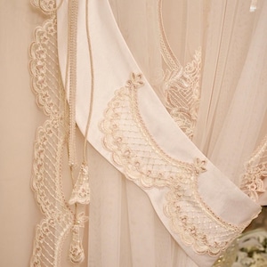 Può includere: Tenda color crema con intricati dettagli in pizzo e un fermatenda decorativo. Il fermatenda presenta un design a nappe e cordoncino, aggiungendo un tocco di eleganza. Il tessuto trasparente della tenda lascia filtrare la luce.