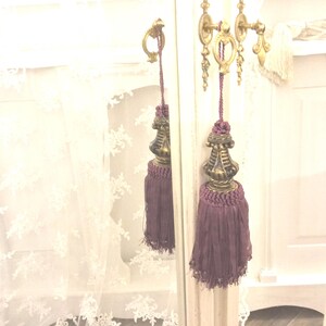 May include: A pair of purple tassel tiebacks with gold metal finials. The tiebacks are hanging from a white door with a gold metal handle.