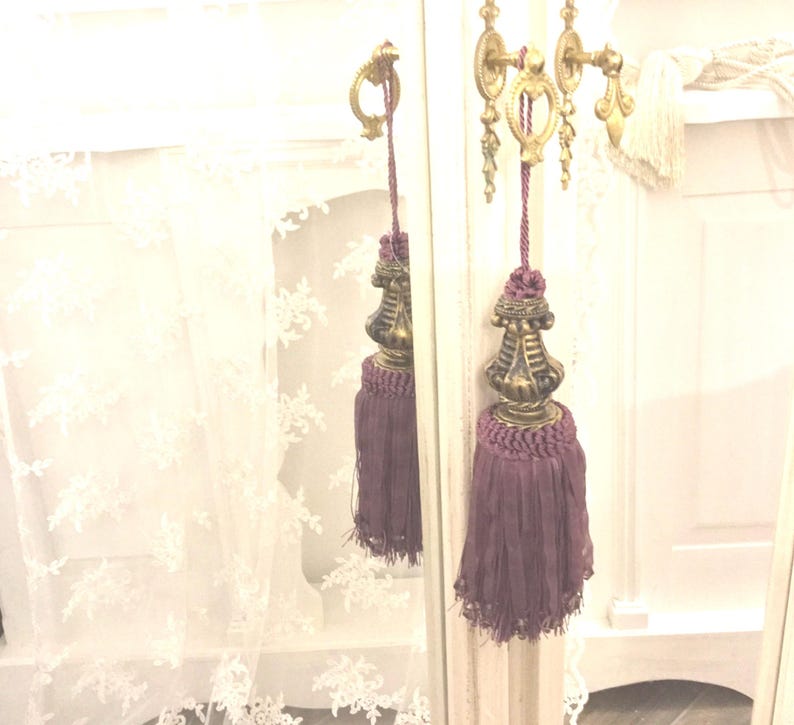 May include: A pair of purple tassel tiebacks with gold metal finials. The tiebacks are hanging from a white door with a gold metal handle.