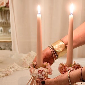 May include: Two lit, pale pink taper candles in decorative holders. The holders are decorated with small pink flowers and ribbons. The candles are held by hands, with a gold cuff bracelet and other bracelets visible.