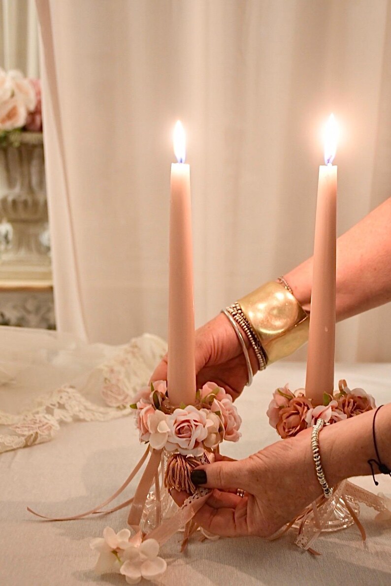 May include: Two lit, pale pink taper candles in decorative holders. The holders are decorated with small pink flowers and ribbons. The candles are held by hands, with a gold cuff bracelet and other bracelets visible.