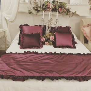 May include: A white bed with a burgundy bedspread and matching pillows. The bedspread has a ruffled edge. The pillows are also ruffled and have a satin finish. There are flowers on the bed.