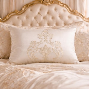 Può includere: Cuscino decorativo color crema con un motivo floreale ricamato, appoggiato su un letto con biancheria da letto abbinata. La testiera è ornata con una finitura dorata. Il tema generale è il lusso e l'eleganza.