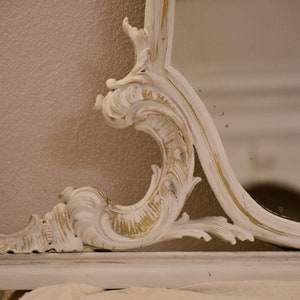 May include: A white ornate mirror with a gold-painted finish. The mirror has a decorative, carved frame with intricate details.