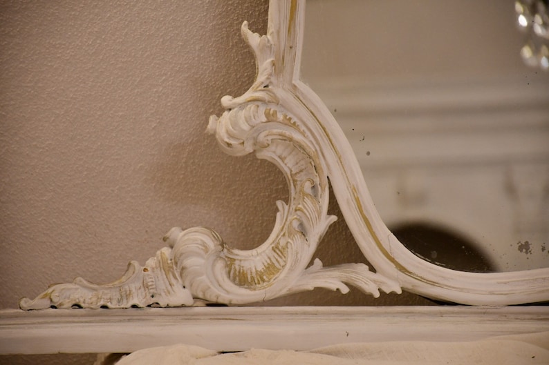 May include: A white ornate mirror with a gold-painted finish. The mirror has a decorative, carved frame with intricate details.