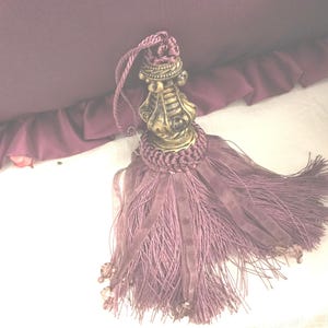 May include: A purple tassel with a decorative gold metal top. The tassel is made of a soft, silky material and has a fringe-like appearance.