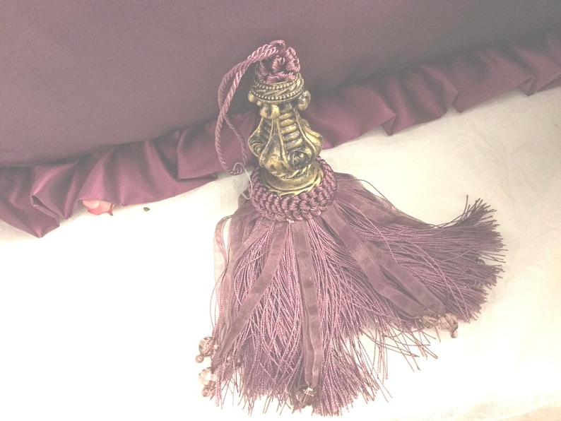 May include: A purple tassel with a decorative gold metal top. The tassel is made of a soft, silky material and has a fringe-like appearance.