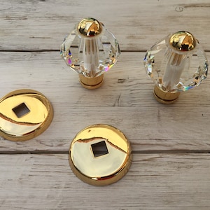 May include: Two clear crystal cabinet knobs with gold accents and two matching gold-coloured backplates with square cutouts. The knobs have a faceted, diamond-like shape. The items are on a wooden surface.