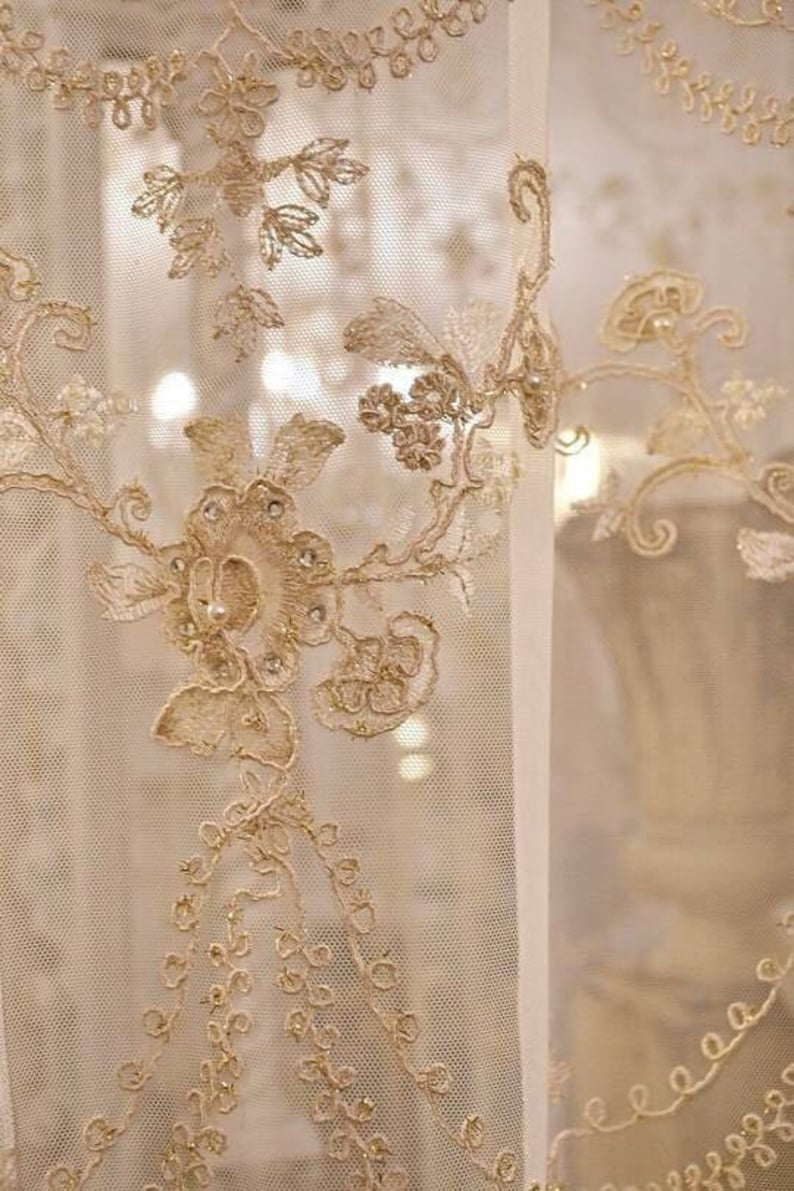 May include: Close-up of a sheer white curtain with delicate gold embroidery. The embroidery features floral patterns and intricate details.