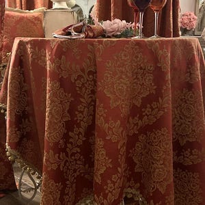May include: A round table covered with a red tablecloth with a gold floral design. The table is set with wine glasses, a floral arrangement, and a decorative napkin. The table has a white metal base.
