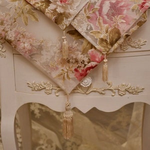 Può includere: Un tavolo bianco con intagli decorativi e un runner da tavolo con motivo floreale e nappe. Il runner è realizzato in tessuto trasparente con fiori rosa e marroni.