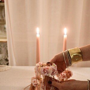 May include: A floral arrangement with pink roses and ribbons is held in front of two lit pink candles. The arrangement is being held by a person wearing a gold cuff bracelet and multiple silver bracelets. The background is a soft pink.