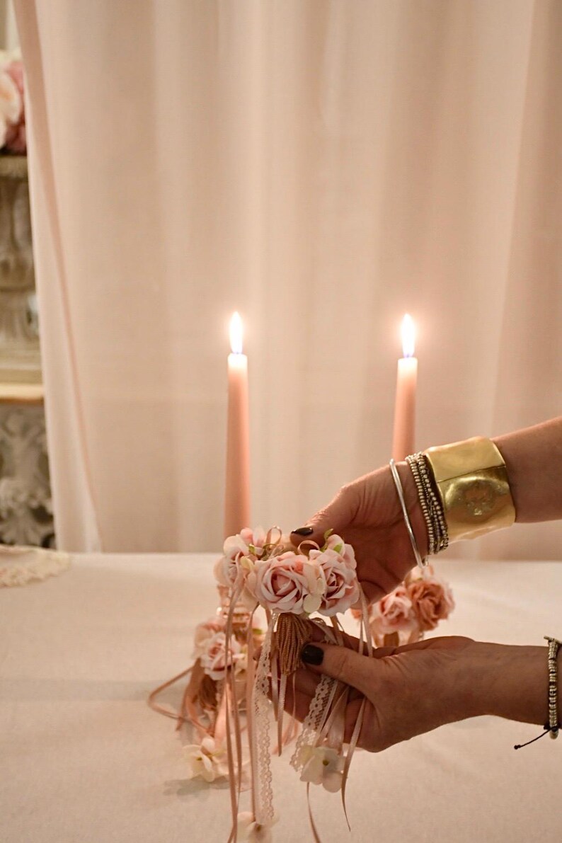 May include: A floral arrangement with pink roses and ribbons is held in front of two lit pink candles. The arrangement is being held by a person wearing a gold cuff bracelet and multiple silver bracelets. The background is a soft pink.