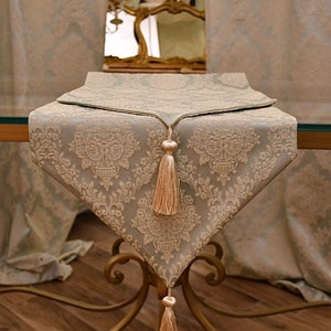 May include: A light blue table runner with a white damask pattern and a beige tassel on each end. The runner is on a glass-top table with a gold metal base.