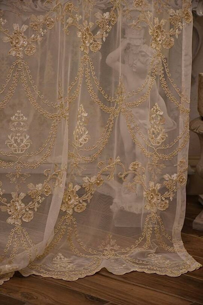 May include: A sheer white curtain with delicate gold embroidery and beaded accents. The curtain has a scalloped bottom edge and a floral pattern.