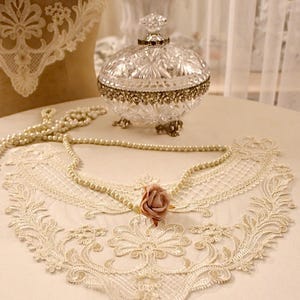 May include: A delicate white lace collar with a pink fabric rose in the center, resting on a white surface with a string of pearls.