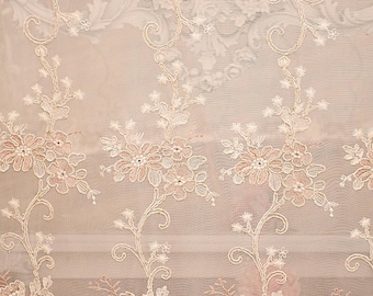 May include: Cream-coloured, sheer lace fabric with a floral design. The pattern features delicate, embroidered flowers and vines in a soft pink hue. The background is a light beige, creating a subtle contrast.