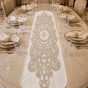 May include: A long, off-white table runner featuring an intricate, symmetrical gold-toned embroidered pattern that runs down the center. The runner has a pointed end finished with a decorative gold tassel, creating a formal table centerpiece.