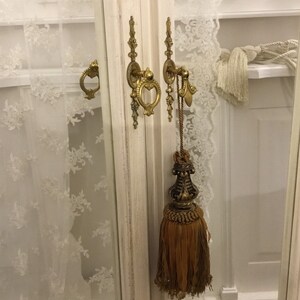 May include: A white cabinet with two glass doors and gold-toned hardware. The cabinet has a decorative tassel hanging from one of the doors.