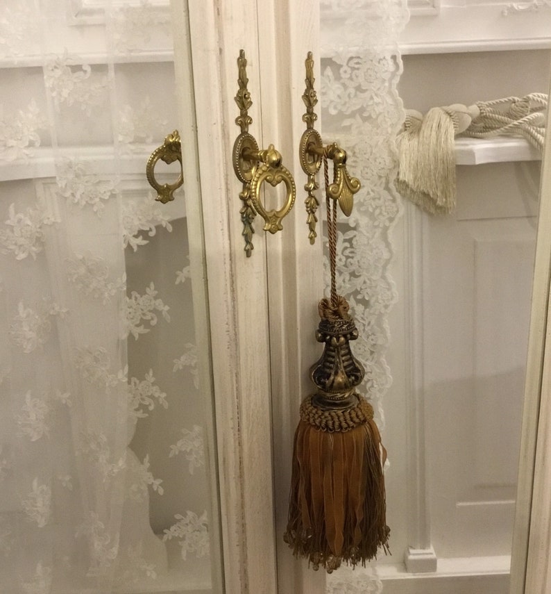 May include: A white cabinet with two glass doors and gold-toned hardware. The cabinet has a decorative tassel hanging from one of the doors.