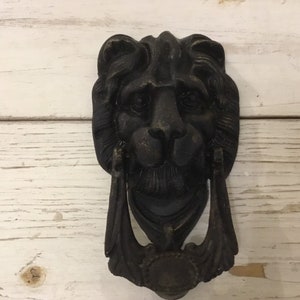 May include: Black metal lion head door knocker with intricate details. The knocker has a ring for knocking and a decorative design around the lion's face.