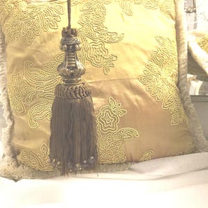 May include: A gold and brown tassel with a decorative metal top. The tassel is hanging from a pillow with an embroidered floral pattern.