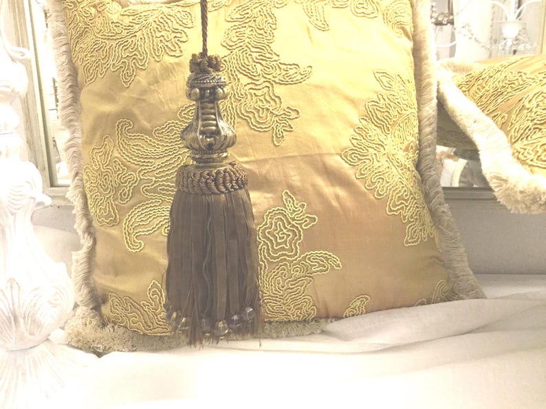 May include: A gold and brown tassel with a decorative metal top. The tassel is hanging from a pillow with an embroidered floral pattern.