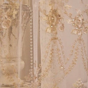 May include: A close-up of a white lace curtain with delicate floral embroidery and a beaded trim. The curtain is draped over a white vase with a textured surface.
