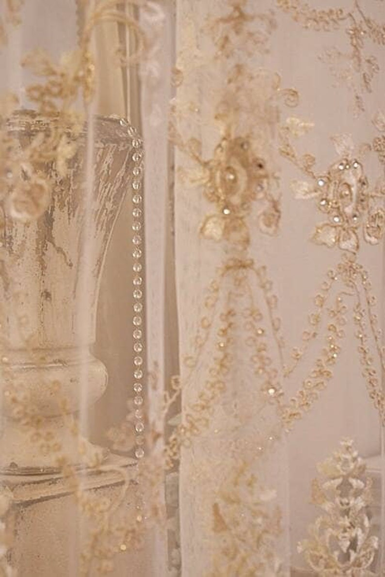 May include: A close-up of a white lace curtain with delicate floral embroidery and a beaded trim. The curtain is draped over a white vase with a textured surface.