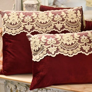 May include: Two burgundy velvet cushions with delicate ivory lace trim. The cushions are arranged on a light wood surface.