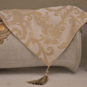 May include: A cream-colored damask table runner with a floral pattern and a tassel. The runner is draped over a white chest with gold accents. The overall theme is home decor.